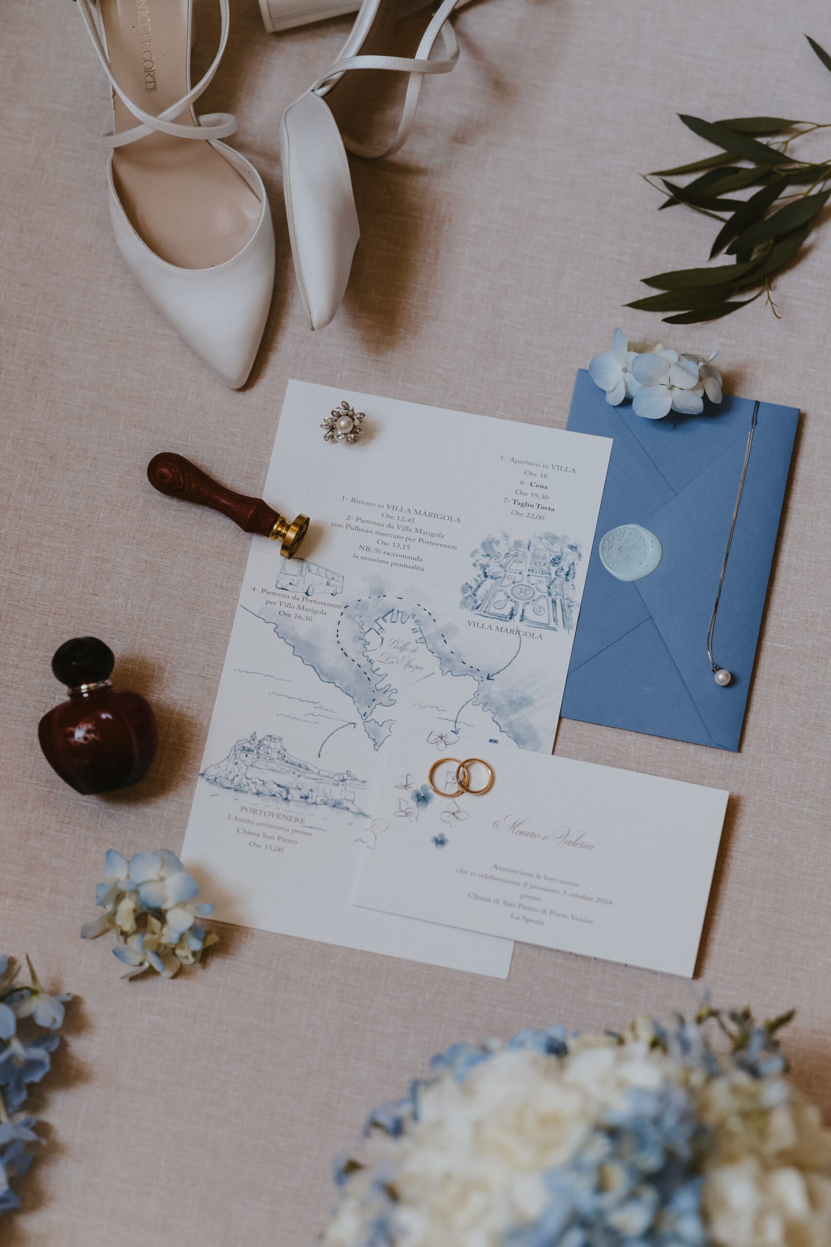Inviti di nozze personalizzati con mappa acquerellata, busta blu con sigillo in ceralacca, scarpe da sposa e anelli nuziali disposti su sfondo in lino chiaro