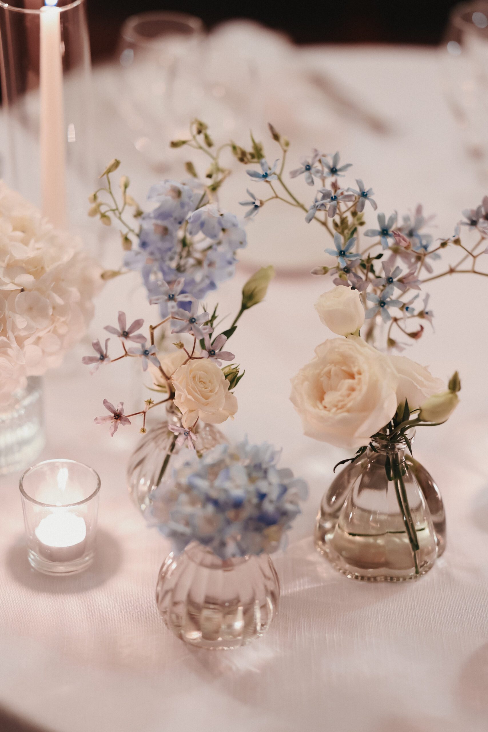 Centrotavola da matrimonio con ortensie azzurre e rose avorio in piccoli vasi di vetro, decorazione elegante con candele per nozze raffinate