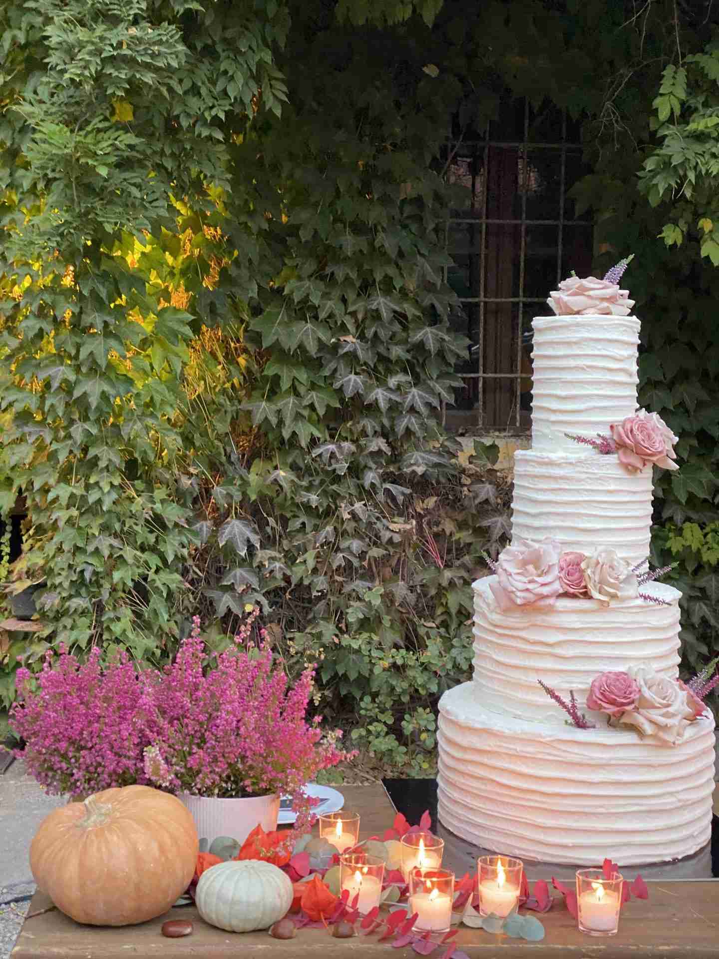 Torta nuziale elegante decorata con fiori autunnali, zucche e candele su sfondo naturale di edera