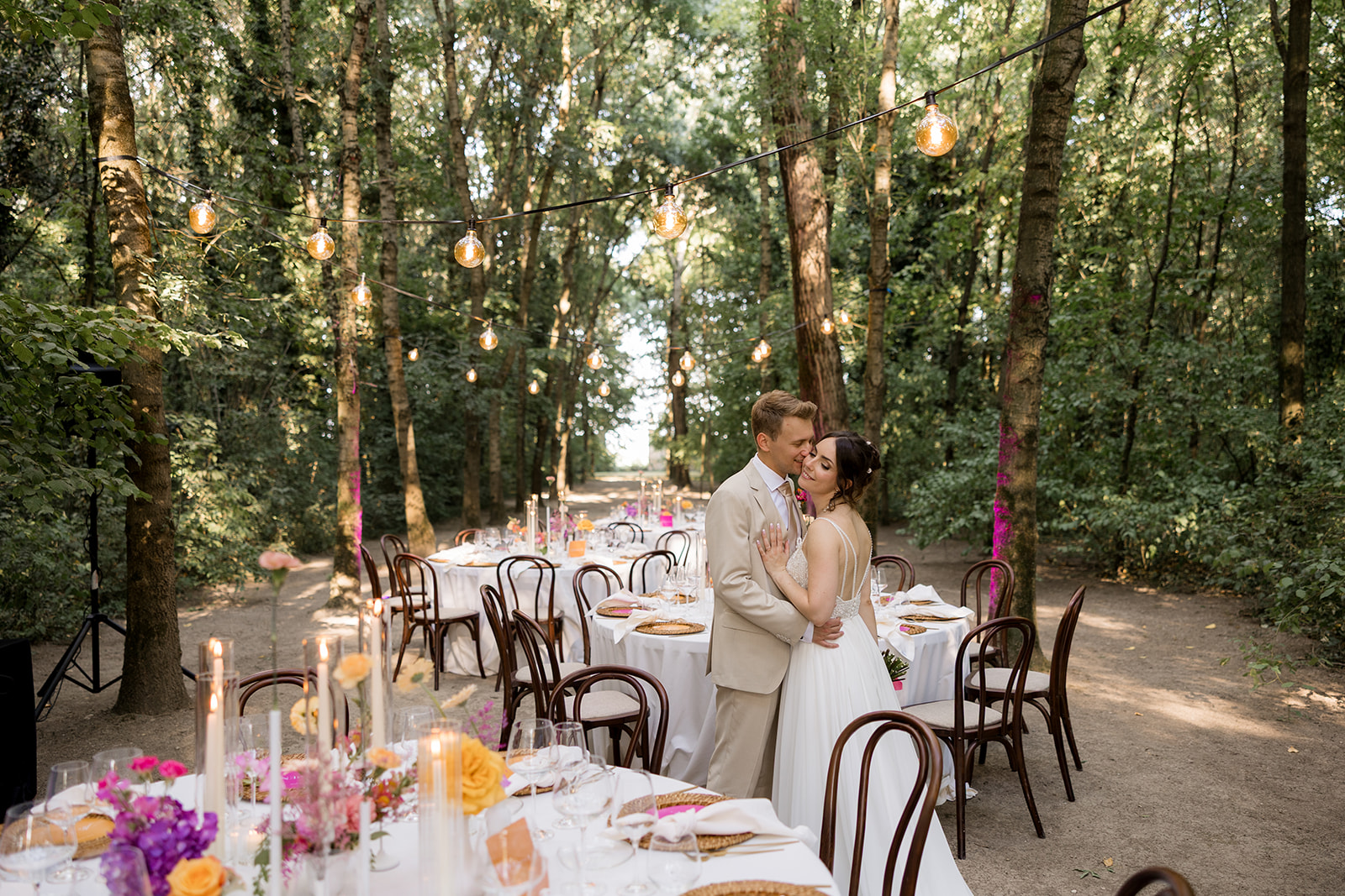 Matrimonio intimo di lusso al Convento dell'Annunciata di Medole — sposi abbracciati nel bosco tra i tavoli della cena, luci sospese e fiori fucsia e arancio